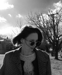 A woman looking down and to her left with some trees behind her, with the wind blowing her hair
