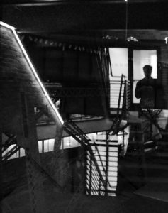 A photograph out a window from a stairwell leads to a high-contrast jumble of lines and angles. Outside the window, you can see overhead rail supports, an elevated train track, and the blur of cars and a truck on an even-higher-up overpass. The interior lights from the stairwell paint contrasting lines onto the window, with the gaps between railings adding grids of light to the center of the frame, and a diagonal light cutting across the left side. Finally, the photographer is lit from behind, reflected in the window, near the top right.