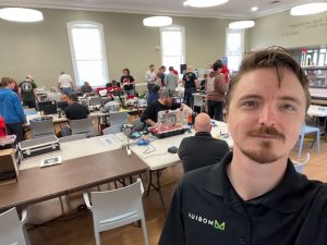 I'm in the foreground of the shot with a single strand of hair draped across my forehead.  There's folks in the background of the shot sitting at or standing around tables, where they're preparing robots to battle.