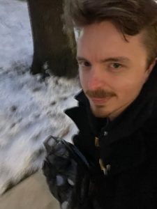 A blurry picture of a man with his tongue slightly out.  There's snow on the ground, and he's holding a bag with water bottles in it.