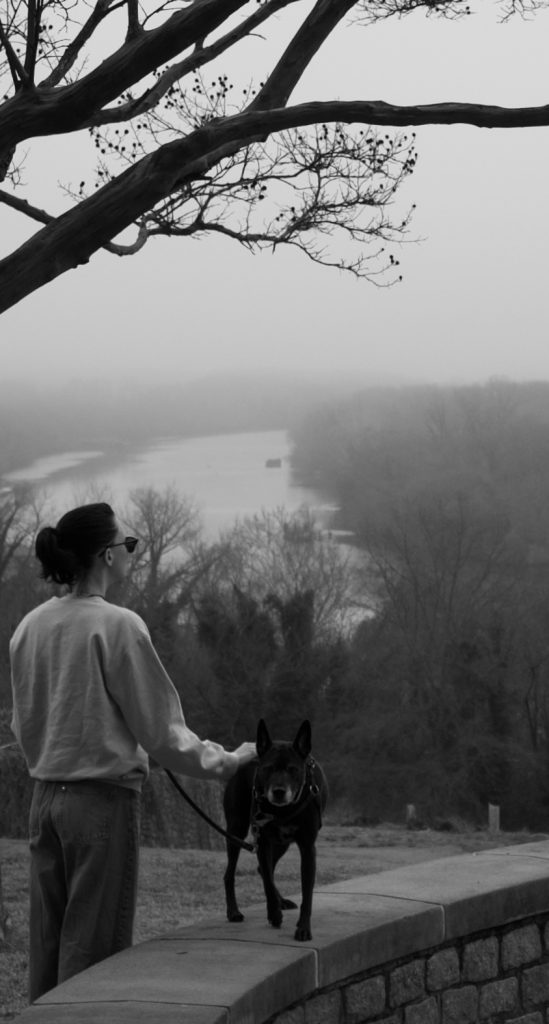 Lana the dog stands on a short wall, looking at the camera with one paw raised. M has one hand on the dog, with her back turned to the camera. The James river lurks in the background.