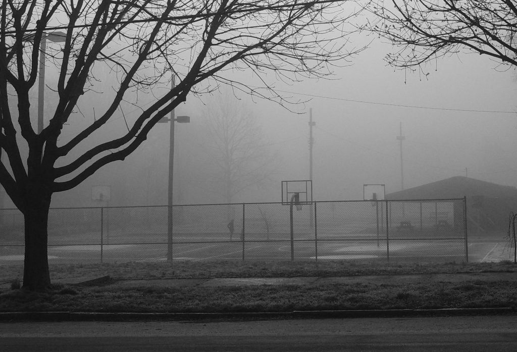 A figure on the basketball courts looks down at a small child, partially obscured by fog.