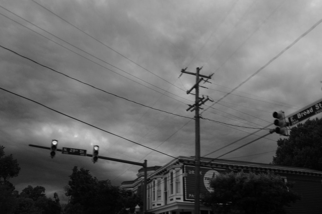 A kind-of blurry greyscale image of power lines, stoplights, and stormy skies above the building Honey Baked Bee is in.
