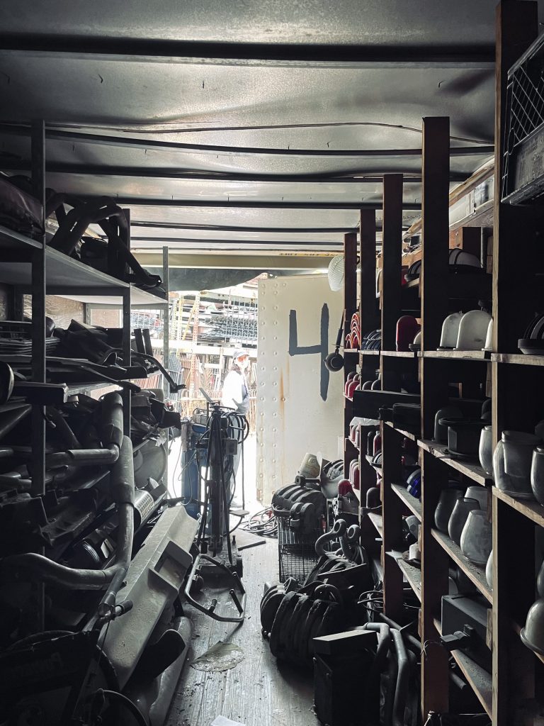The inside of an architectural surplus container, with chaotic shelves of pipes and lighting fixtures, and a big "4" painted on a door in the background.